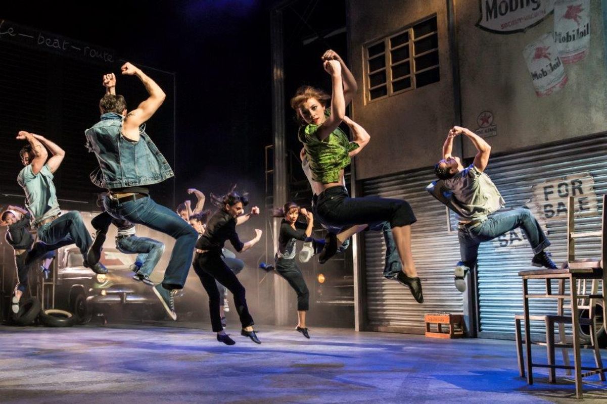 Performers energetically leap in unison on stage in a dynamic dance scene, set against an urban backdrop with graffiti, exuding excitement and passion.
