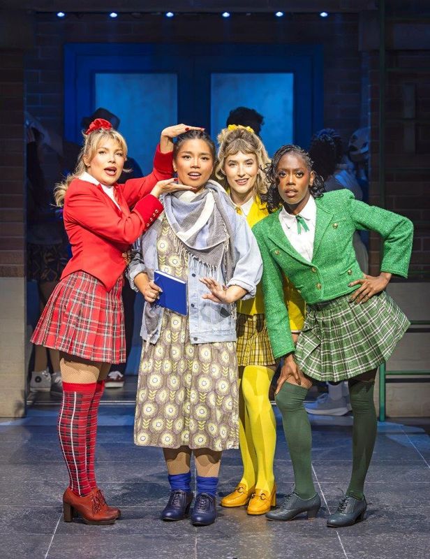 Four women stand in a row on stage. Three wear colorful outfits: a red jacket and plaid skirt, green jacket and skirt, and yellow jacket and checkered skirt. They pose confidently around a woman in muted colors, conveying a mix of dominance and unease in a theatrical setting.
