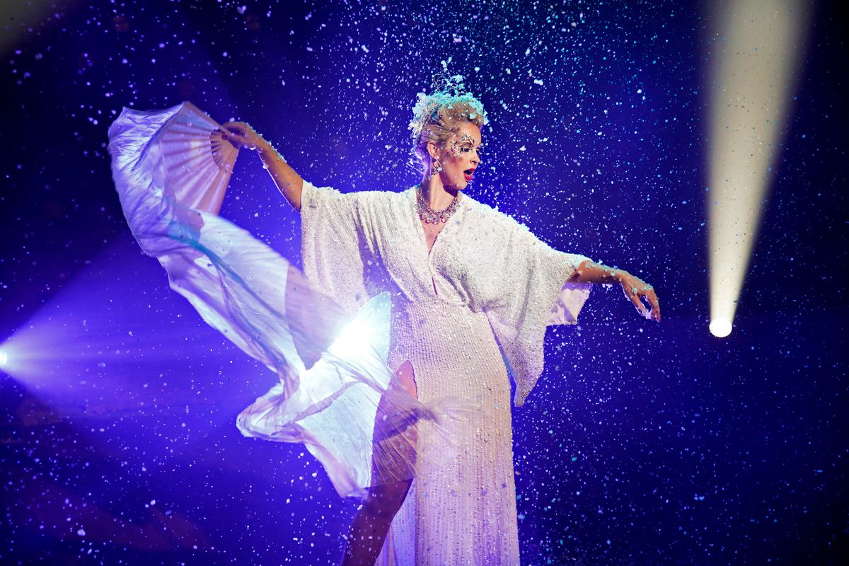 A performer stands wearing a white sequined dress holding a white fan.