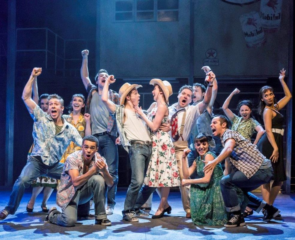 Energetic group of performers in vintage outfits, mid-dance onstage. Two central figures in hats and floral attire embrace joyfully, surrounded by cheering cast.
