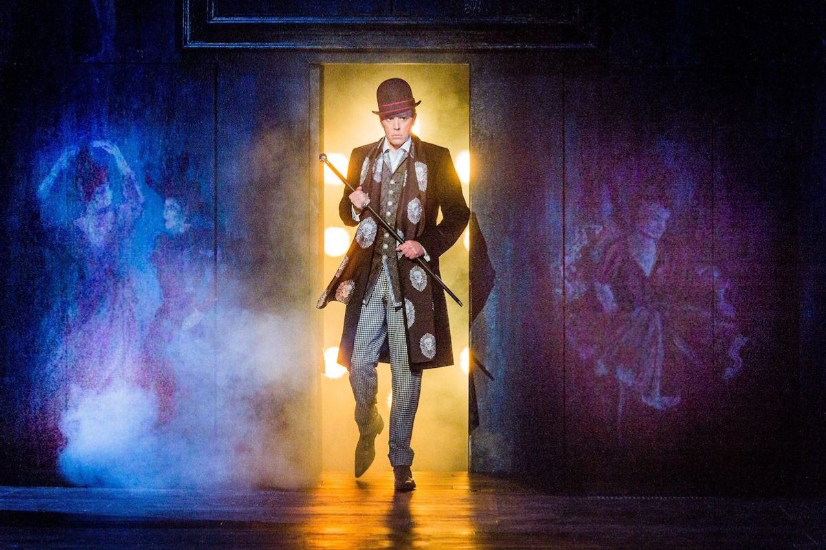 A person wearing a bowler hat, suit and holding a cane is walking out of a lit room