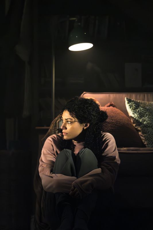 A woman sits on a dimly lit floor, leaning against a sofa. She looks contemplative and pensive, illuminated by a hanging lamp, creating a moody ambiance.