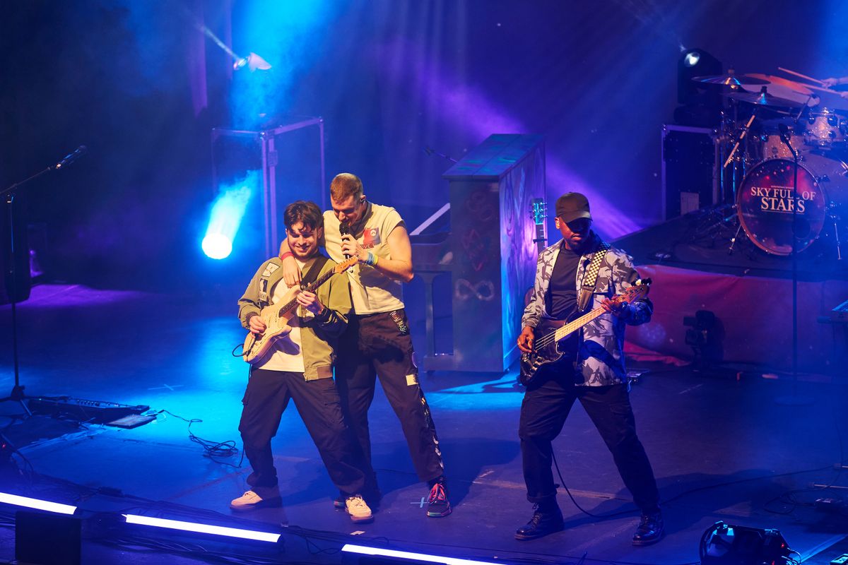 A band on stage with vibrant blue and purple lighting. The lead singer engages the crowd, flanked by energetic guitarists. The mood is lively and dynamic.