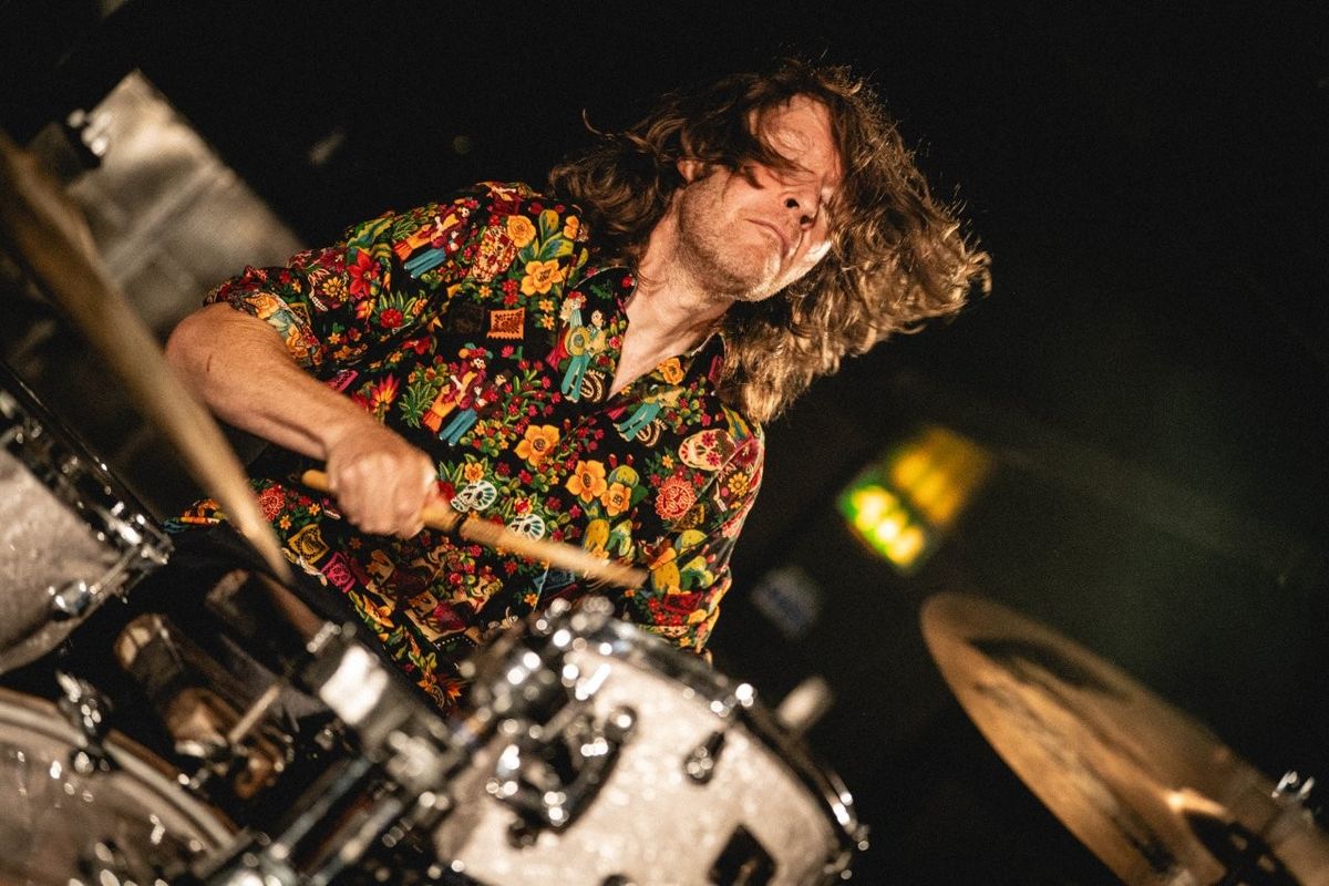 A person energetically plays a drum set, captured mid-action. They are wearing a colorful, patterned shirt and have long, flowing hair. The environment is dimly lit, suggesting a live performance atmosphere.