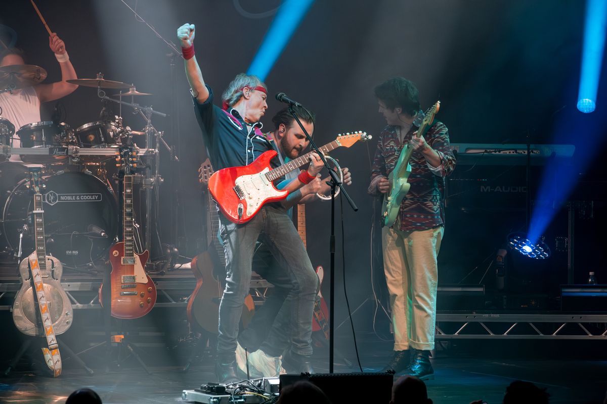 Band on stage performing, they're playing guitars and drums and there are flashing lights around them