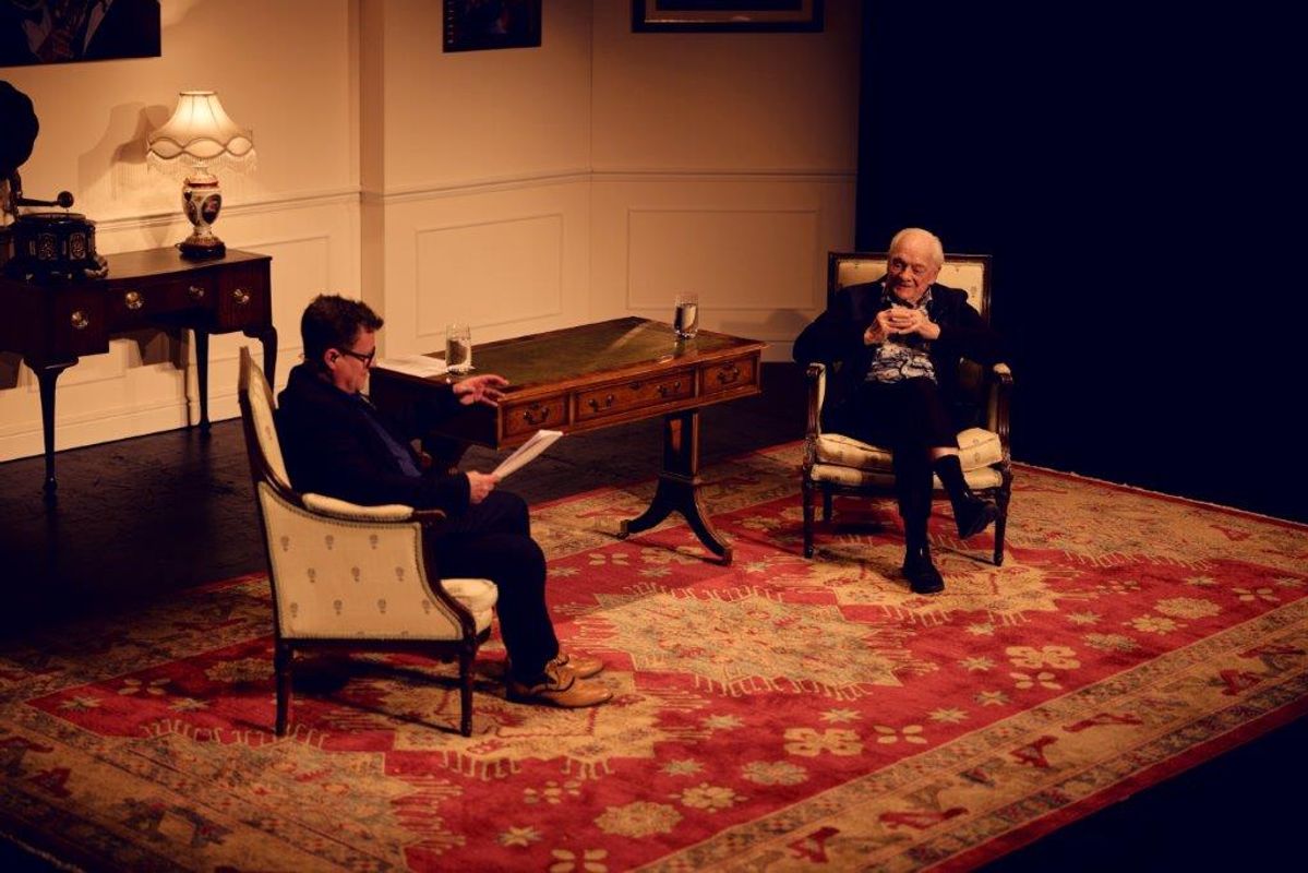Sir David Jason on stage with another gentleman. The setting is made of two chairs, a dark wooden desk and a large rug