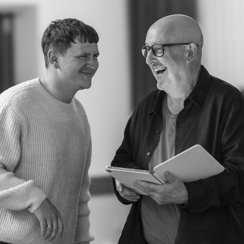 Two men are looking toward each other, one is holding a script