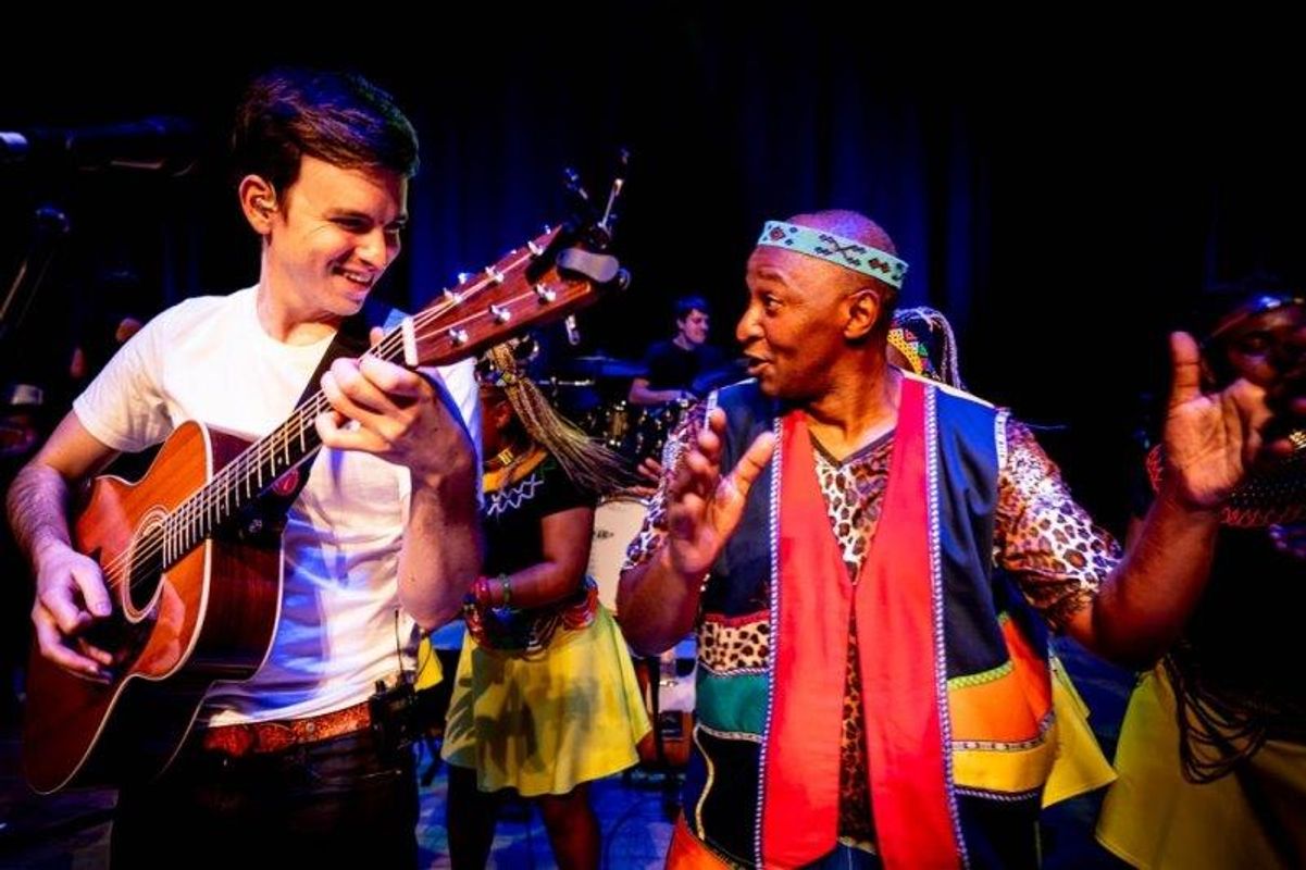 The South African Cultural Choir are singing on stage with Paul Simon. They are all wearing bright and vibrant African clothing. Paul is playing a guitar