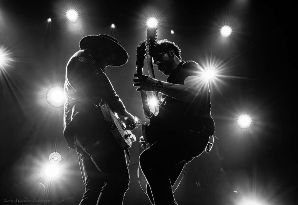 Two musicians are performing on stage, playing electric guitars. The image is in black and white, with bright stage lights creating a dramatic silhouette effect. The musicians appear to be interacting energetically during their performance.