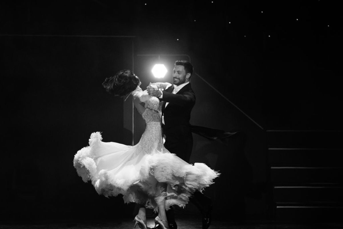 Giovanni and a woman dancer, he is wearing a black suit and white shirt. The woman is wearing a flowing elegant white dress