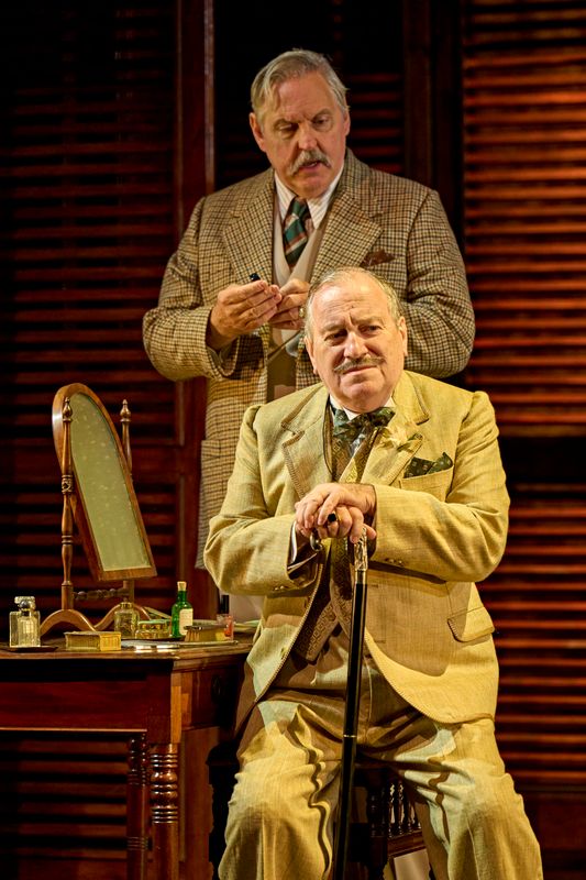 A person with a walking stick sits down in front of someone wearing a checked suit.