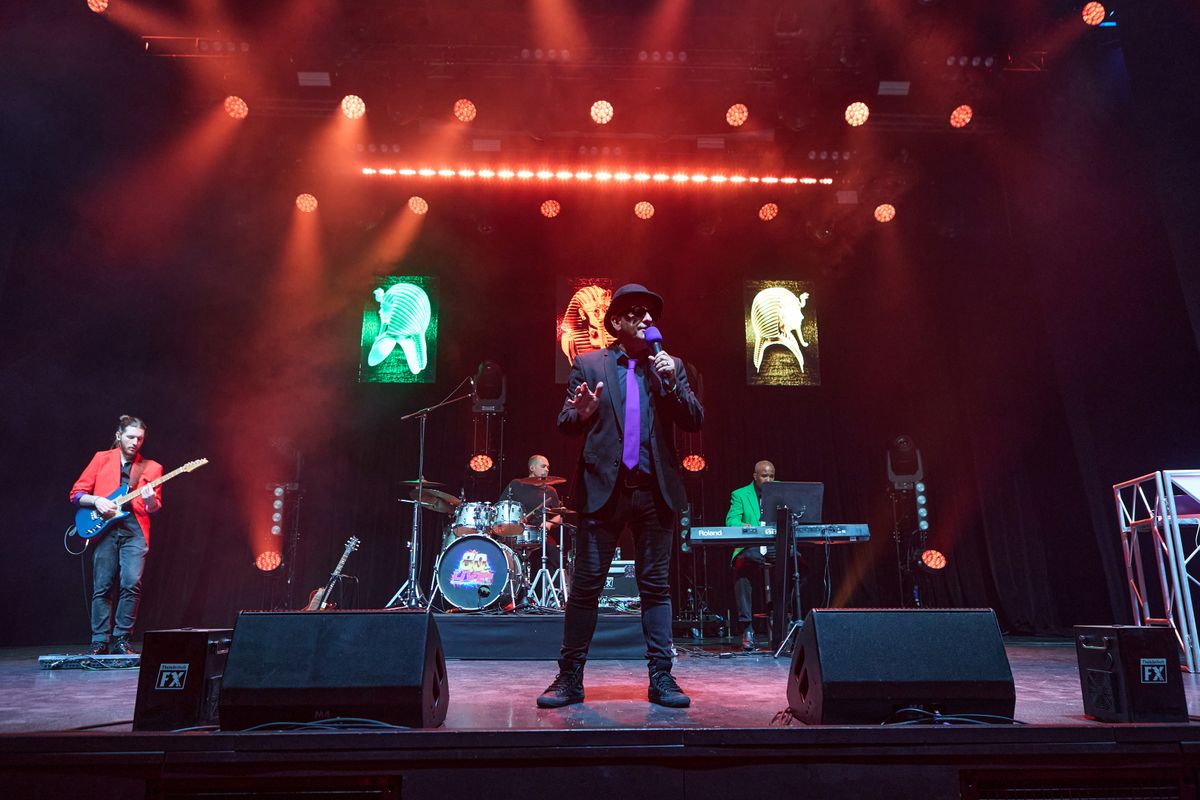 A person wearing a black blazer and purple tie sings on stage.