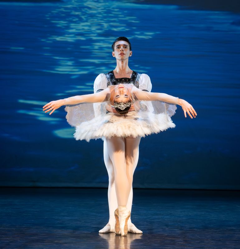 Two ballet dancers stand on stage with the dancer in front draped backwards.