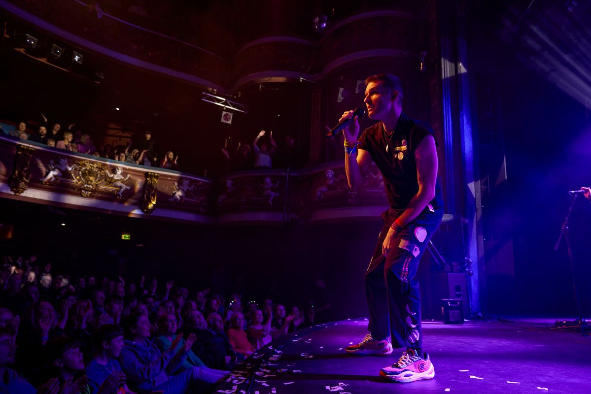 A performer on stage sings into a microphone, surrounded by vibrant purple lighting. An enthusiastic audience claps and cheers in a dimly-lit theater.