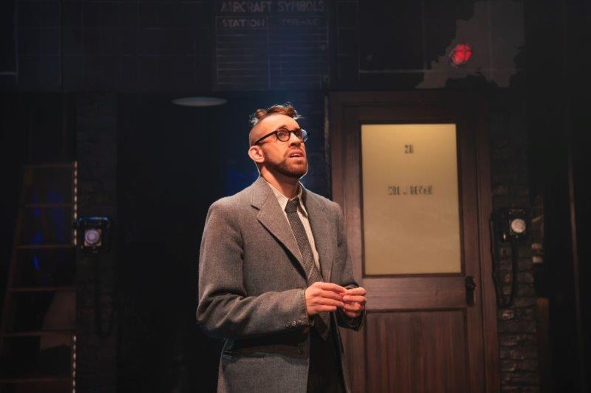 A man in a vintage suit stands on stage, looking up thoughtfully. The background features a wooden door with frosted glass and dim stage lighting.