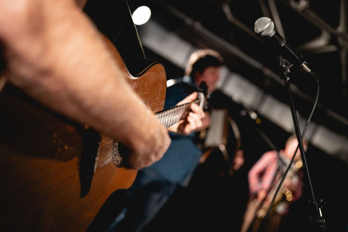 Musicians performing on stage, with a focus on a person playing a guitar in the foreground. Other musicians, slightly blurred in the background, include one with an accordion and another with a saxophone. A microphone is visible.