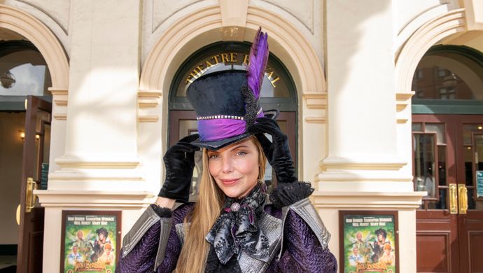 An actor playing the villain poses in black velvet gloves and a black top hat adorned with a purple feather.