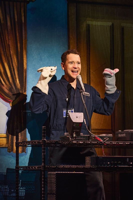 Daniel stands in front of a microphone with a sock puppet on each hand.