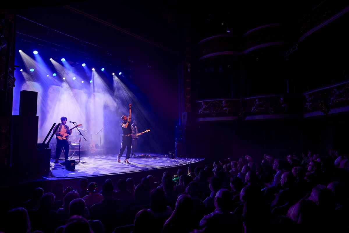 A lively concert scene in a dimly lit theater. A band performs energetically on stage under blue spotlights, with a cheering audience in the foreground.