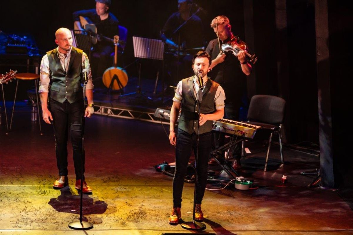 Two male singers perform on stage in vests and shirts, with a violinist and guitarist in the background. The mood is energetic and engaging.