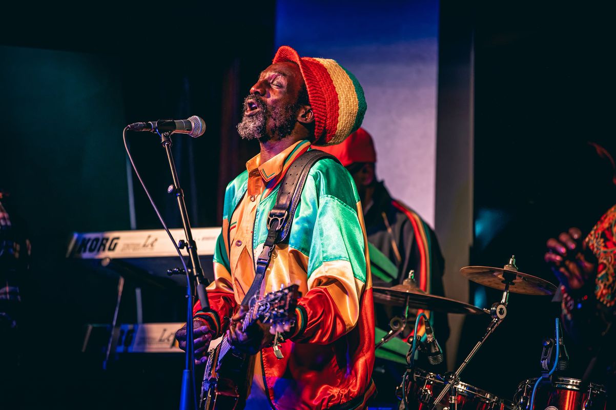 A musician passionately plays guitar on stage, wearing a colorful Rastafarian jacket and hat. A drum set and keyboard are visible in the background.