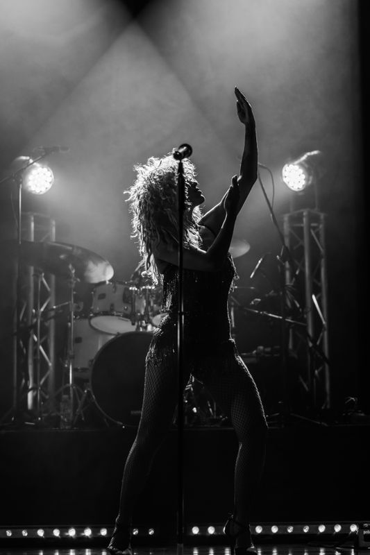 A person lit by the spotlight looks to the left hand side with both arms raised 