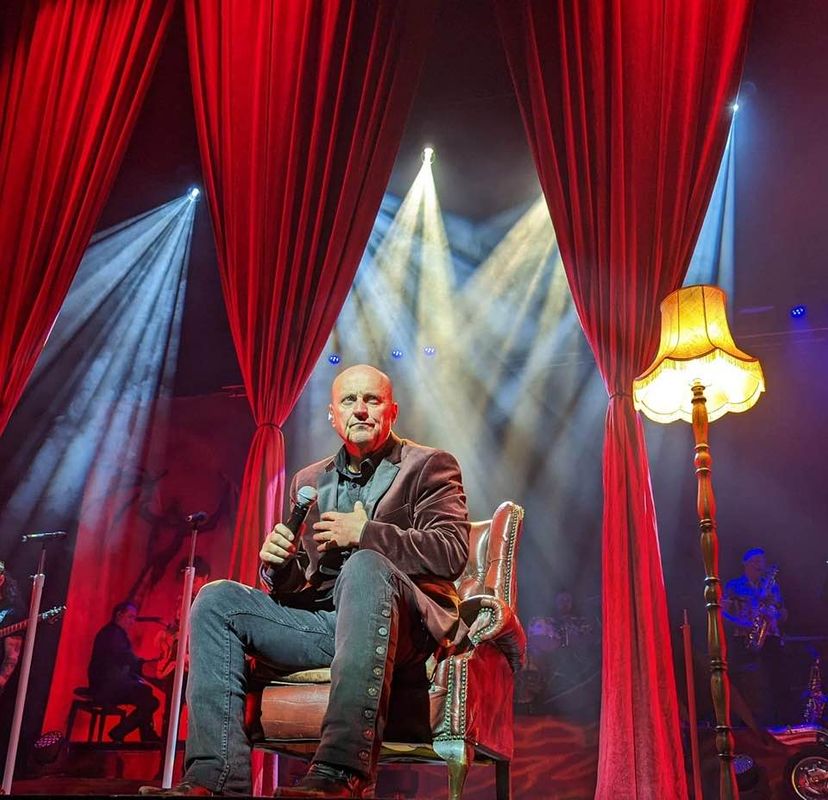 Steve Steinman holds a microphone while sitting on a leather chair onstage, surrounded by dramatic red curtains and warm lighting. Musicians play in the dimly lit background.