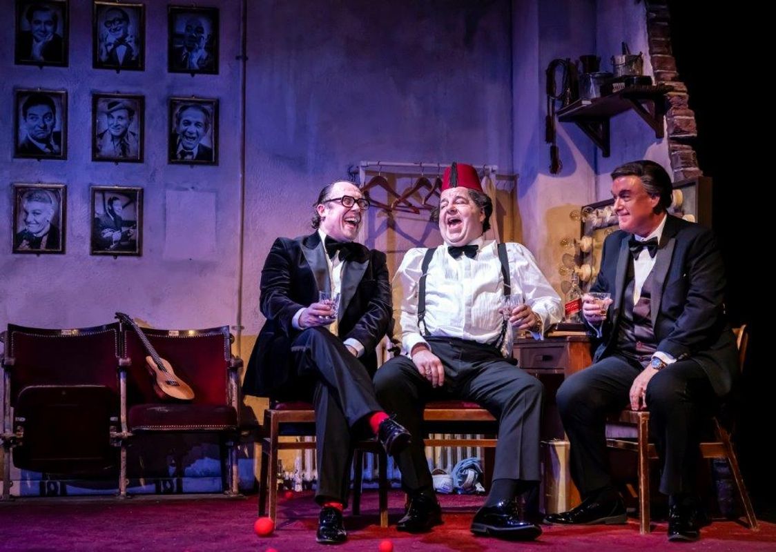 Three men in tuxedos sit on stage, laughing and holding drinks. A guitar rests on chairs nearby. The backdrop features framed black-and-white portraits.