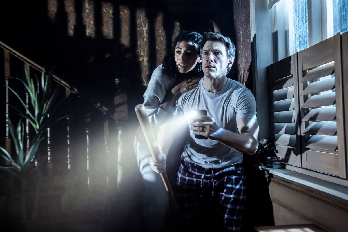 A man in pajamas holds a flashlight and a bat, looking anxious, with a woman clutching his arm. They stand in a dimly lit room near shuttered windows.