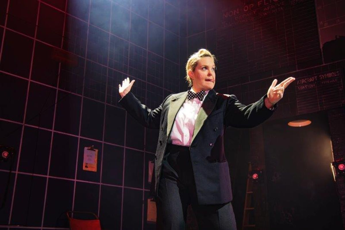 A performer in a formal suit and bowtie stands confidently on stage, arms raised. The background features grid patterns and dim stage lighting, creating a dramatic tone.