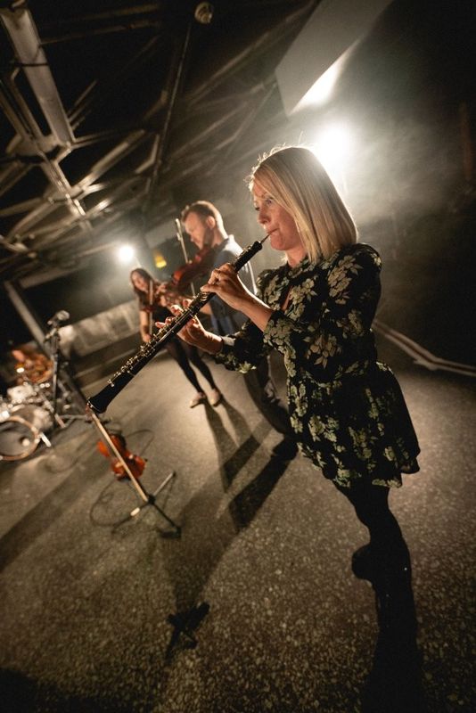 Musicians perform on a dimly lit stage. One plays a violin, while another plays a flute. Additional instruments, including a drum set and a microphone, are visible in the background. The setting has industrial-style lighting.