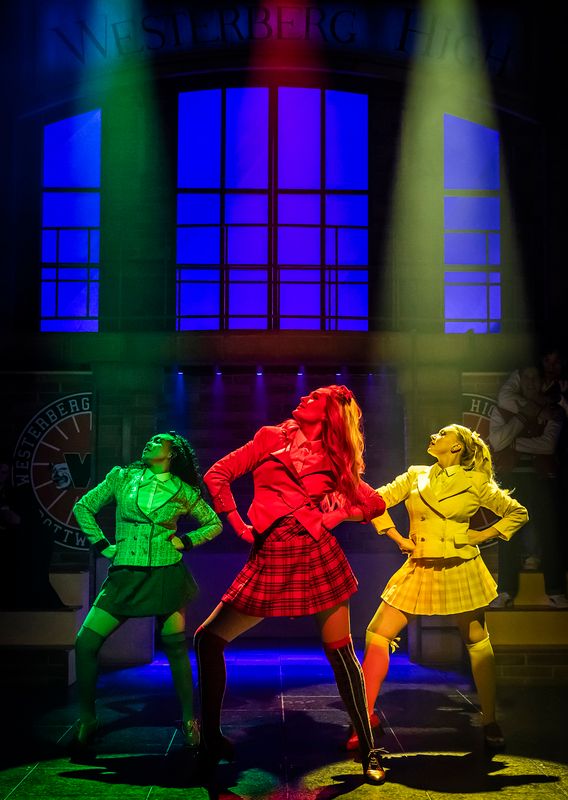 Three performers pose confidently on stage, wearing bright 1980s-style outfits: green, red, and yellow. Dramatic blue backlighting and colored spotlights add intensity.