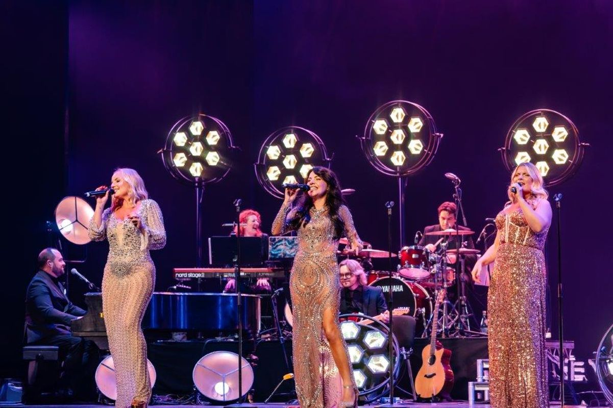  Kerry Ellis, Louise Dearman and Rachel Tucker in sparkling gowns sing on stage, backed by a live band with illuminated circular lights. The scene is vibrant and energetic.