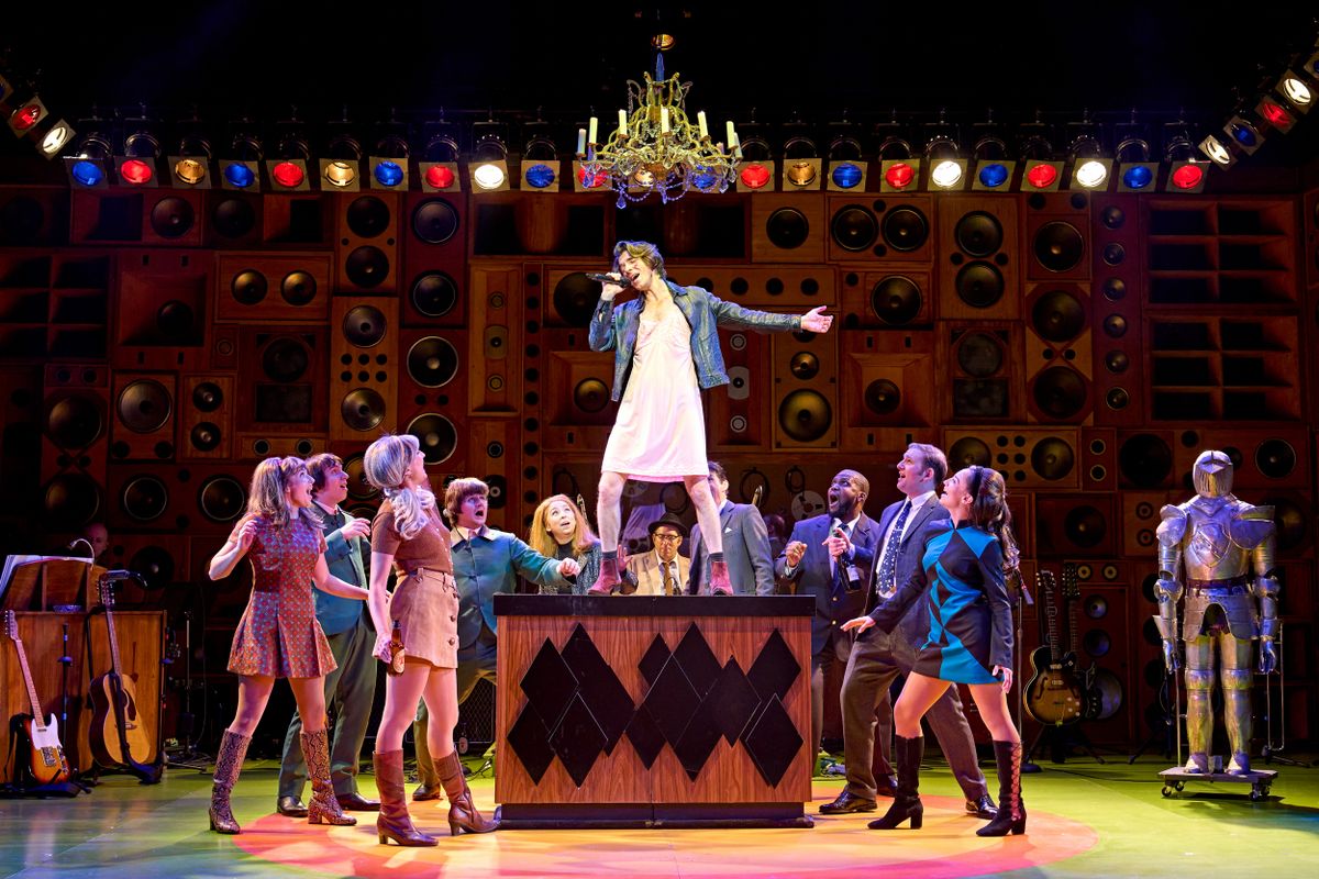 A person wearing a dress and denim jacket stands on a box with the surrounding ensemble looking shocked.