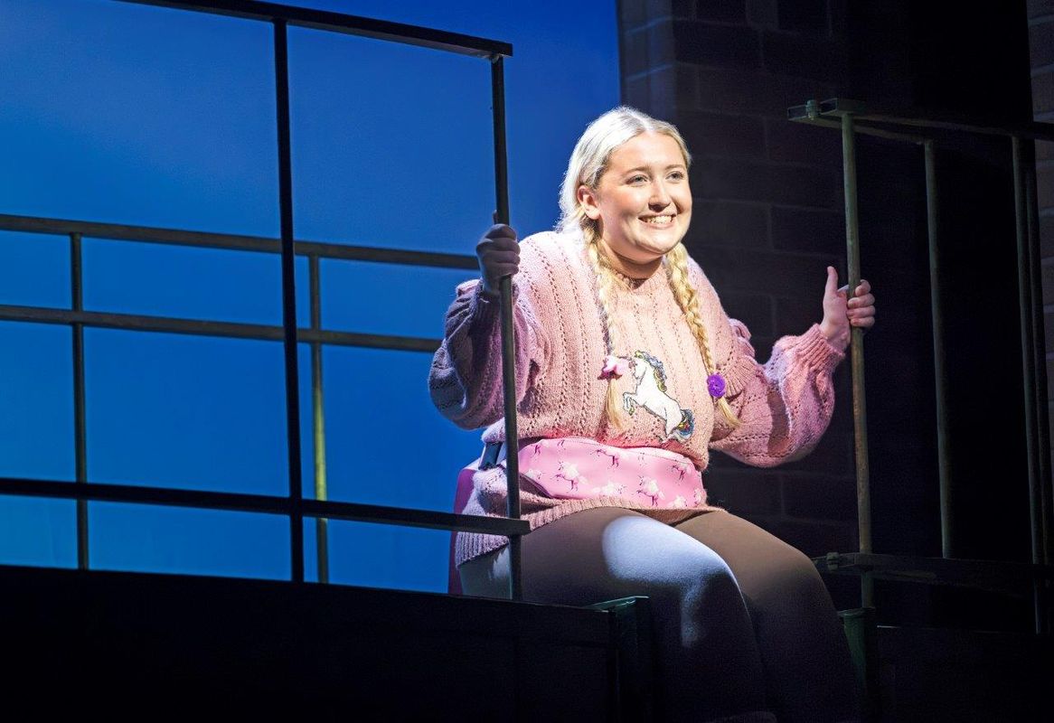 A girl with blonde hair with two braids and is wearing a pink jumper with a unicorn on it. She is sitting on a ledge and is smiling on.