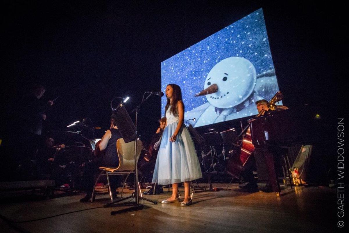 People performing on stage with The Snowman on a projector behind them.