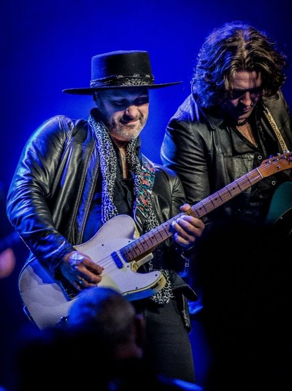 An individual playing an electric guitar on stage, wearing a black leather jacket and a wide-brimmed hat, accompanied by another person also playing guitar. The background is illuminated in blue light.