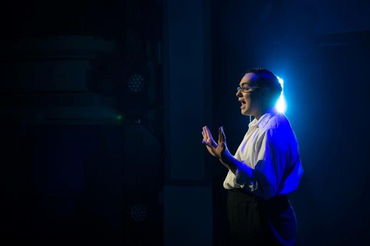 A person in glasses, wearing a white shirt, passionately speaks on stage under dramatic blue lighting, conveying intensity and focus.