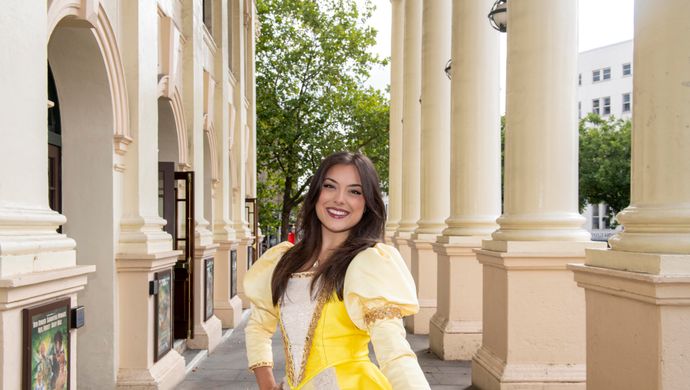 An actor playing the princess stands outside the Theatre Royal, next to the pillars, posing in a yellow dress.