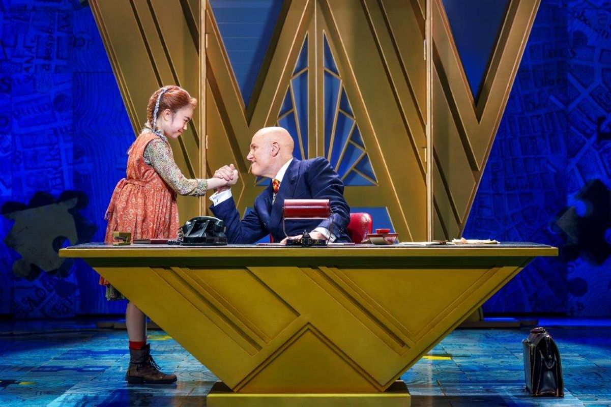 There is a gentleman sat at a large desk, the young girl is arm wrestling with him