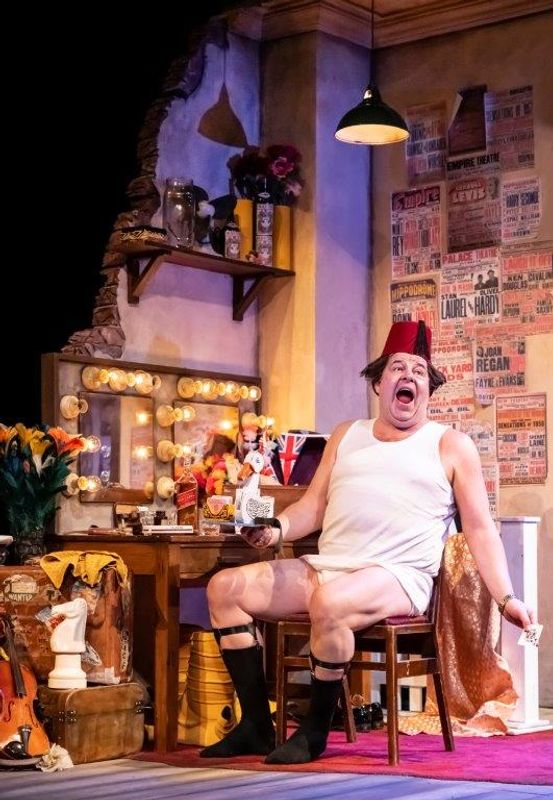 A man in a comedic outfit, wearing a fez and undershirt, sits onstage with an exaggerated expression. The room is cluttered and lively, featuring a vintage vanity with lights, posters, and props. The atmosphere is theatrical and whimsical, evoking humor and nostalgia.