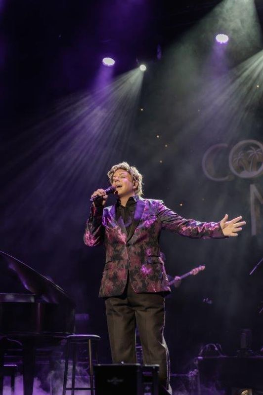 Martyn James in a floral blazer passionately sings on a dimly lit stage, holding a microphone. Purple lights and mist create a dramatic, lively atmosphere.