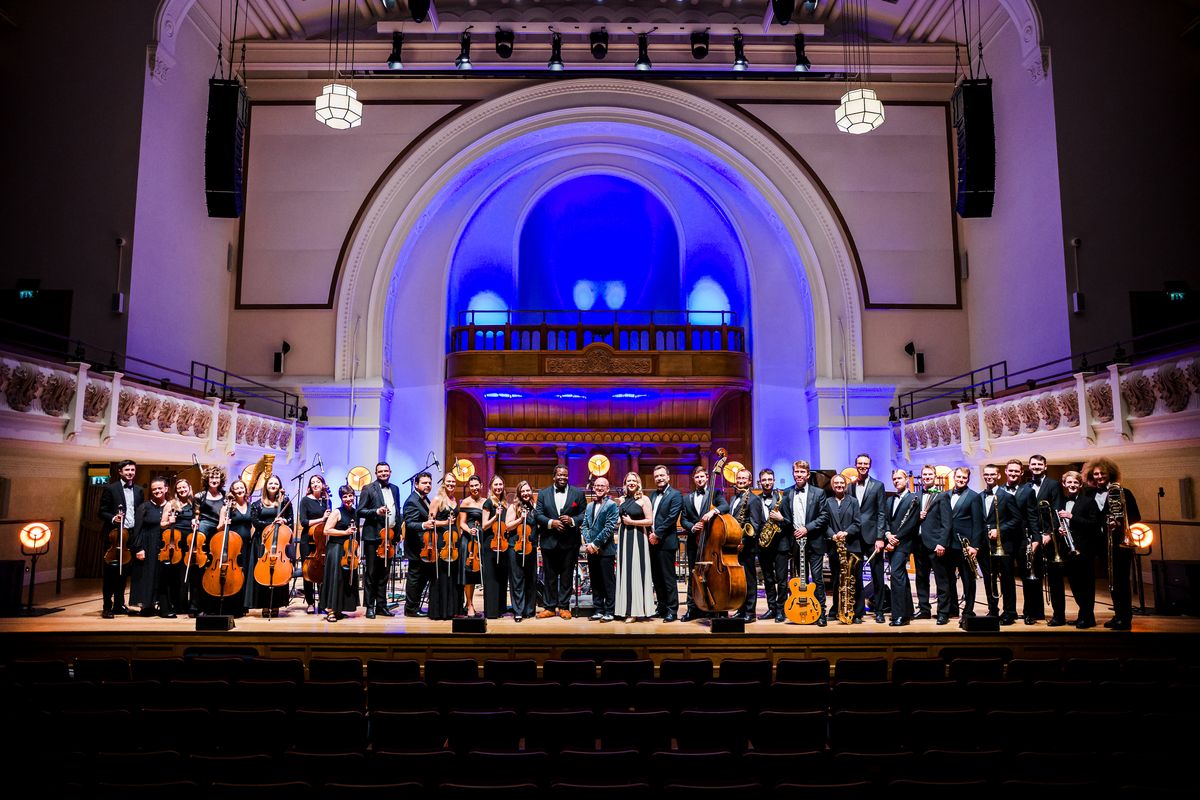 The whole orchestra stand smiling on stage.
