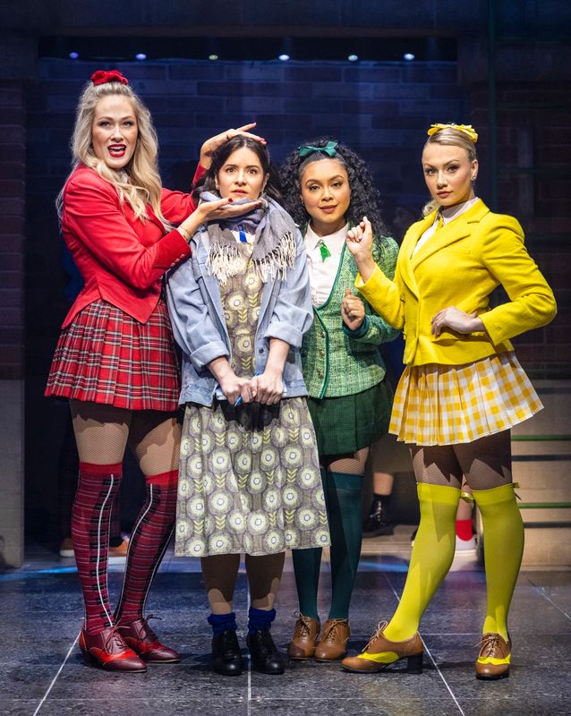 Four women stand in a row on stage. Three wear colorful outfits: a red jacket and plaid skirt, green jacket and skirt, and yellow jacket and checkered skirt. They pose confidently around a woman in muted colors, conveying a mix of dominance and unease in a theatrical setting.