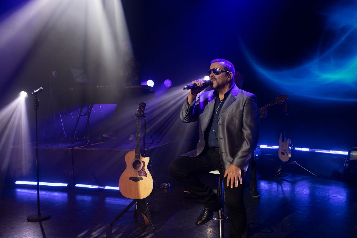A man performing on stage to an audience. There are colourful lights. Behind him are backup singers and a band playing