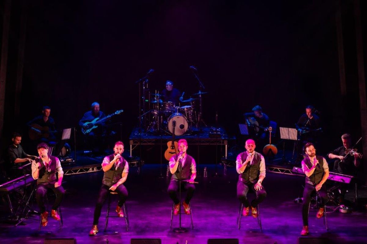 The Shamrocks sit on stools, singing on a dimly lit stage. They are backed by a live band, creating a warm, energetic atmosphere.