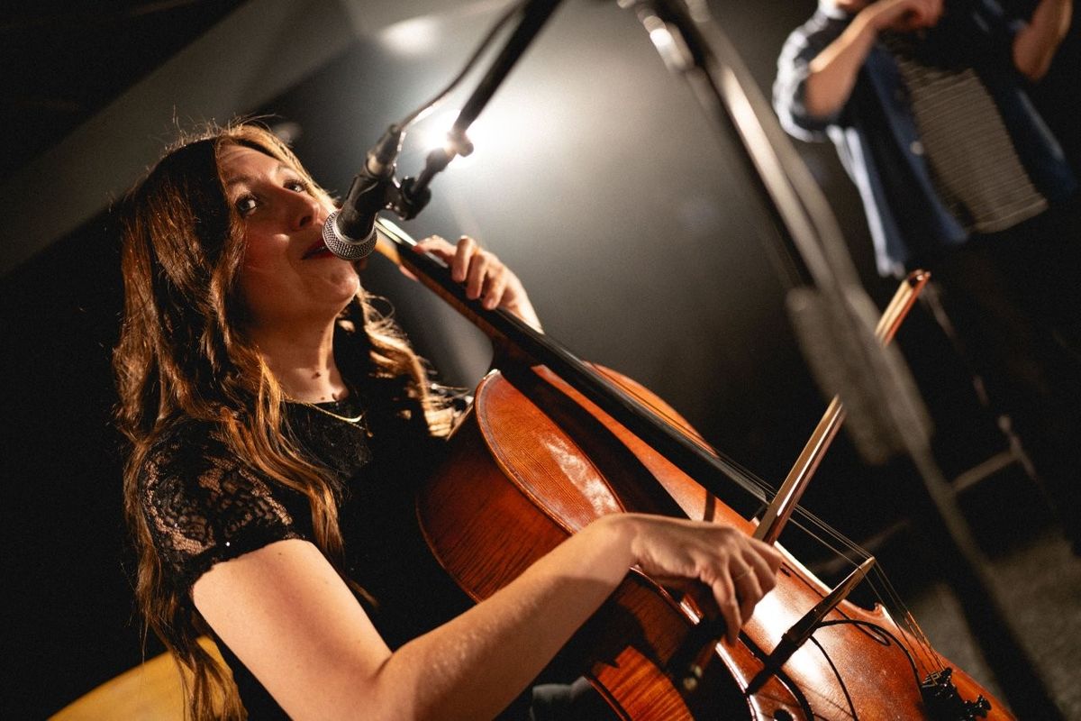 A person is playing the cello and singing into a microphone on stage. The setting is dimly lit, creating a focused atmosphere on the performance.