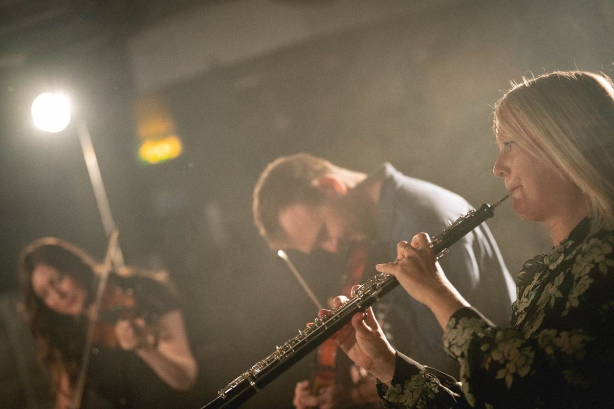 Musicians playing instruments in a dimly lit setting. The person in the foreground plays a clarinet, while others in the background play violins. The focus is on the music performance, with a subtle spotlight creating a warm atmosphere.