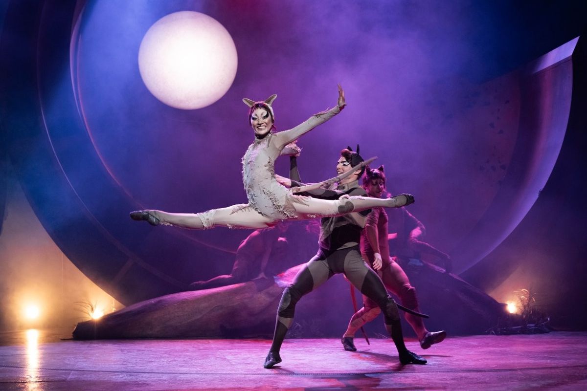 A performer dressed in an elaborate animal-inspired costume poses mid-air during a dance routine on a stage. The background features a large, glowing full moon and atmospheric mist, enhancing the theatrical ambience.