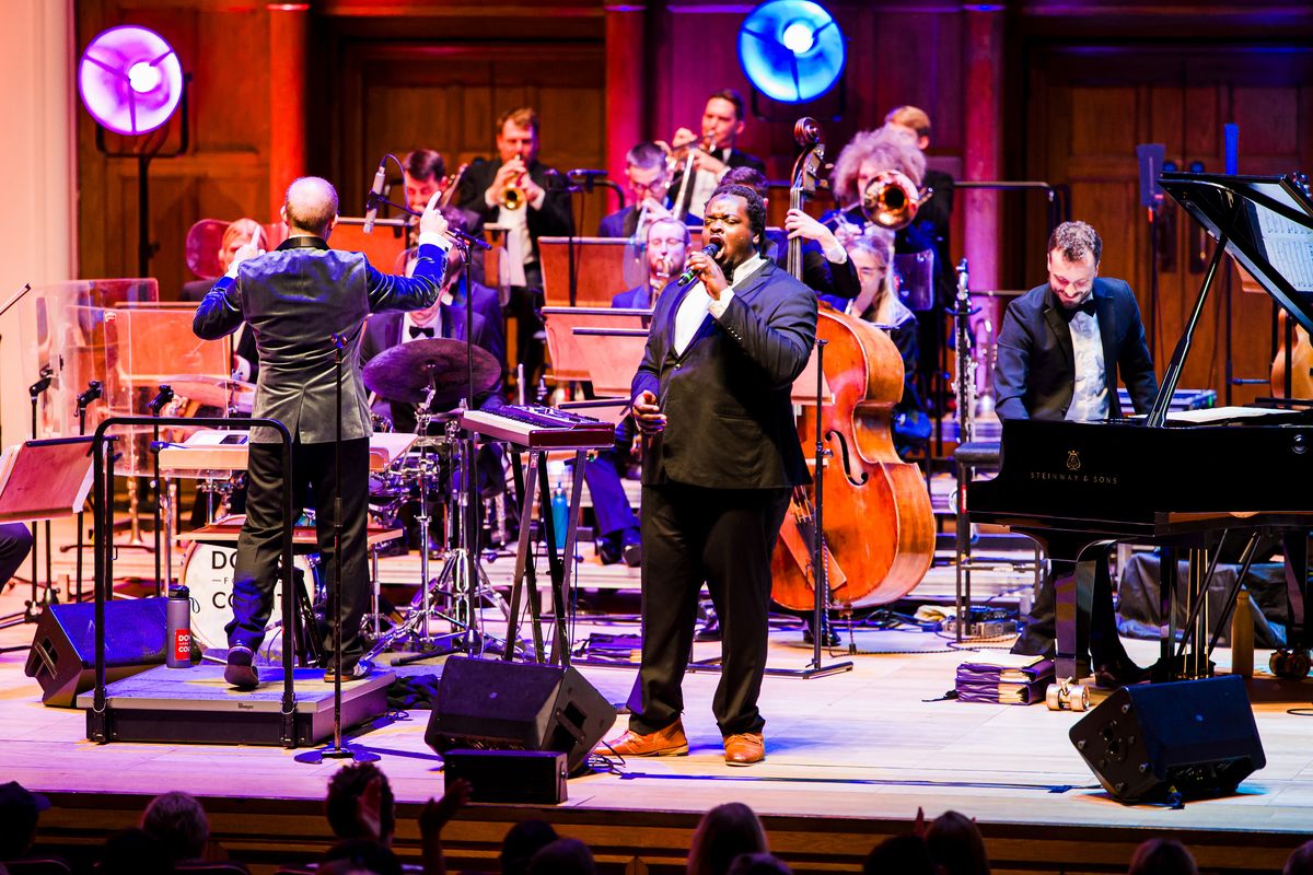 As singer, wearing a dark suit, is accompanied by the orchestra.